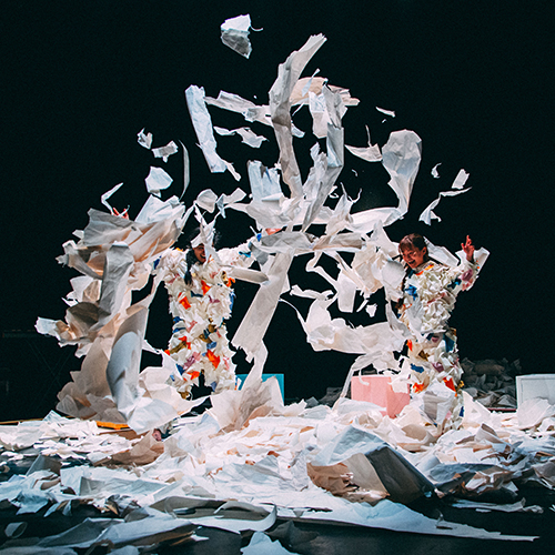 Zwei Personen stehen auf einer Theaterbühne, tragen Anzüge aus Papier und werfen Papierfetzen in die Luft