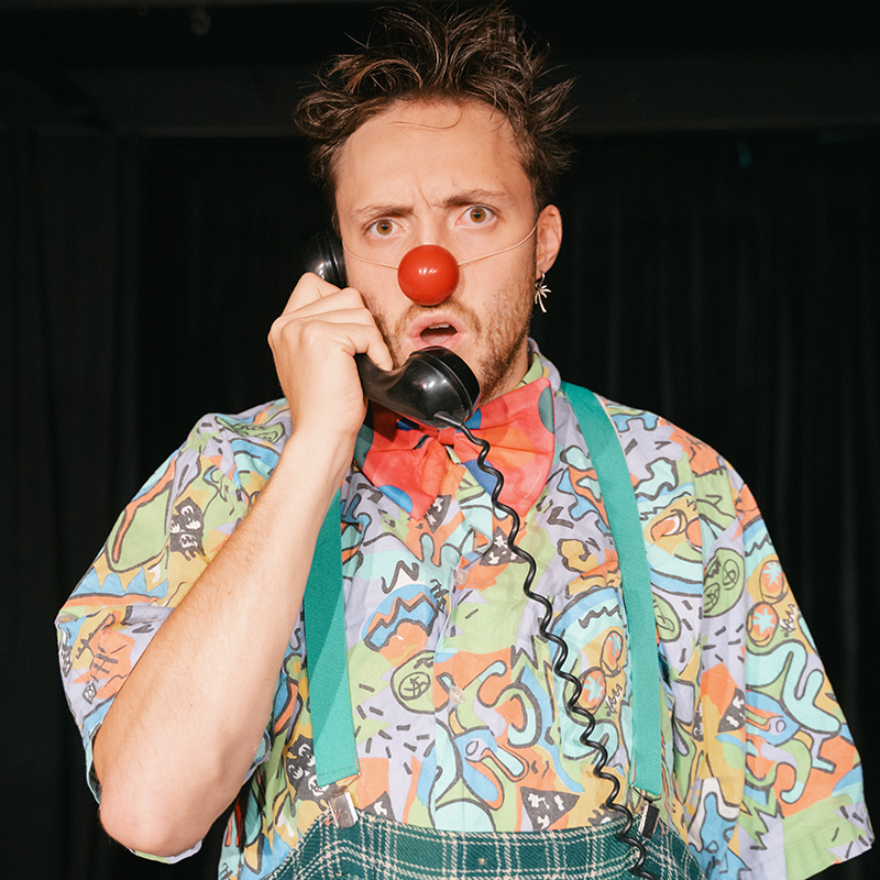 Schauspieler mit Clownsnase, buntem Hemd und Hosenträgern, hält einen Telefonhörer ans Ohr und schaut erstaunt in die Kamera