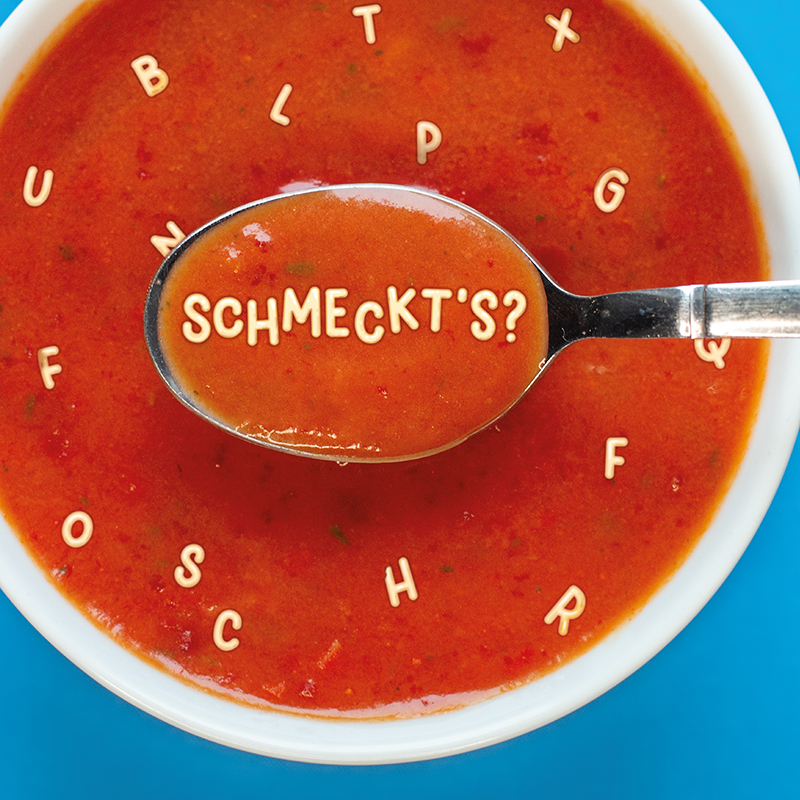 Tomatensuppe mit Buchstaben, darüber ein Löffel in dem aus Buchstaben "Schmeckt’s?" steht