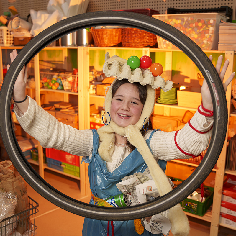 Ein Kind in einem kreativen Kostüm aus recycelten Materialien lächelt und hält einen großen runden Gegenstand vor sein Gesicht, im Hintergrund Regale mit diversen Gegenständen.