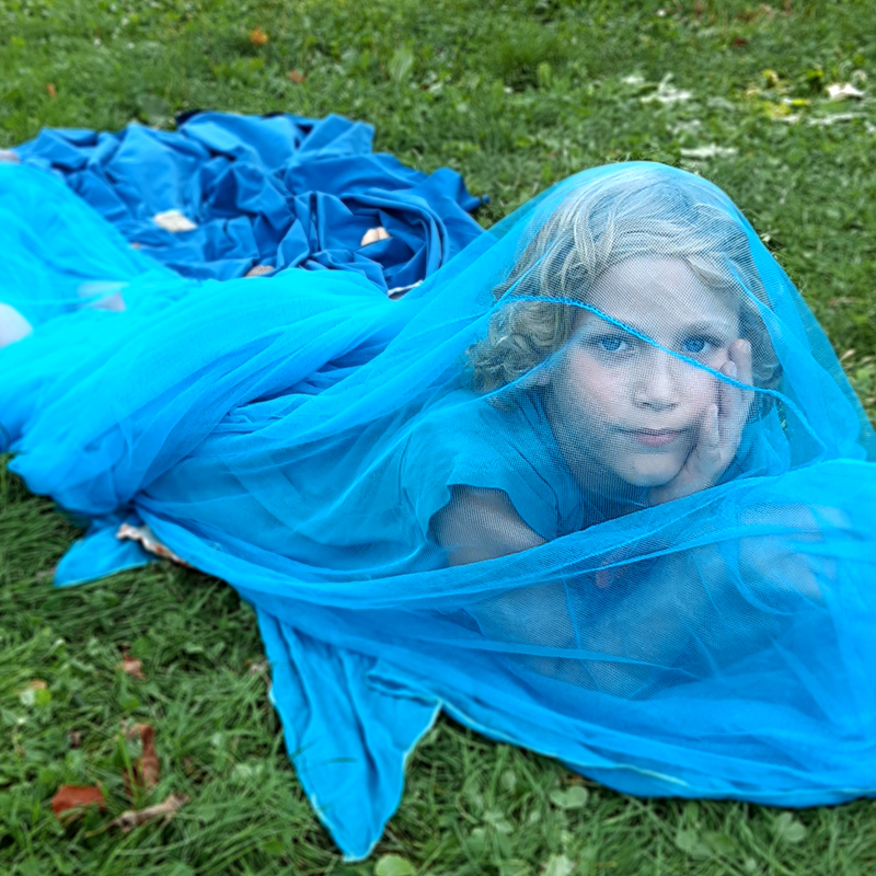 Kind unter einem Tüllstoff, auf einer Wiese liegend