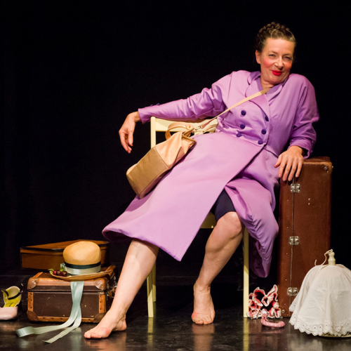 KNOPFTheater_Theater Asou_Rapunzel (c) Dagmar Leis Schauspielerin im Mantel sitzt lässig auf einem Hocker, rund herum Koffer und Gepäck