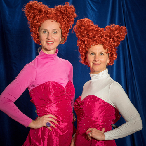 Schauspielerinnen mit roten Locken-Perücken und rosa-Kleidern strahlen in die Kamera