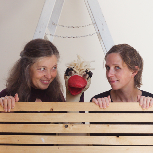 Theaterfoto: zwei Frauen und eine Gans-Handpuppe schauen über eine Bretterwand