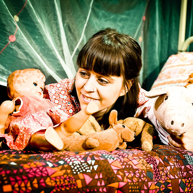 Theaterfoto: Junge Frau liegt mit Puppe und Kuscheltieren auf dem Bett