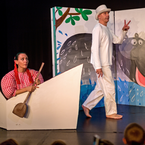 Theaterfoto: Schauspielerin im Kostüm sitzt in einem Papierboot mit Paddel, daneben Schauspieler in weißer Kleidung und weißem Zylinder
