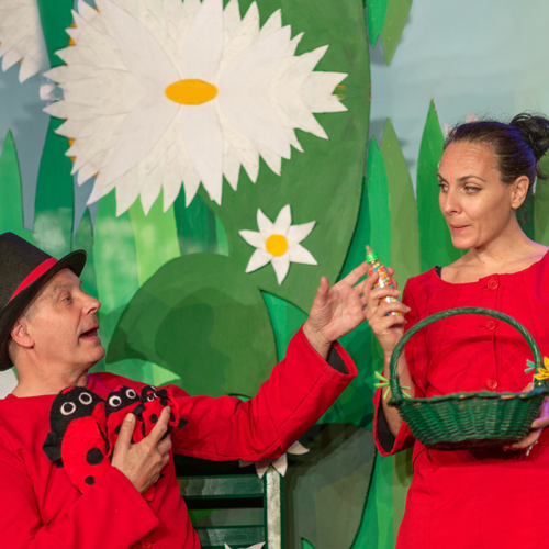 Schauspieler, in rot gekleidet mit zwei Marienkäfer-Kuscheltieren in den Händen, kniet vor rot gekleideter Schauspielerin und greift nach Süßigkeiten