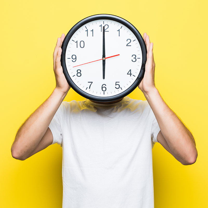 Sommeröffnungszeiten: Mann in weißem Tshirt hält eine große Uhr vor seinem Gesicht, sie zeigt 18 Uhr an