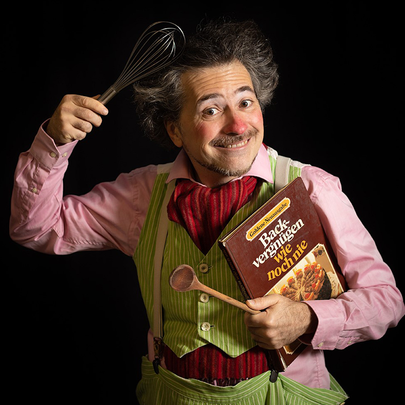 Schauspieler als Clown verkleidet, mit Schneebesen und Kochbuch in den Händen