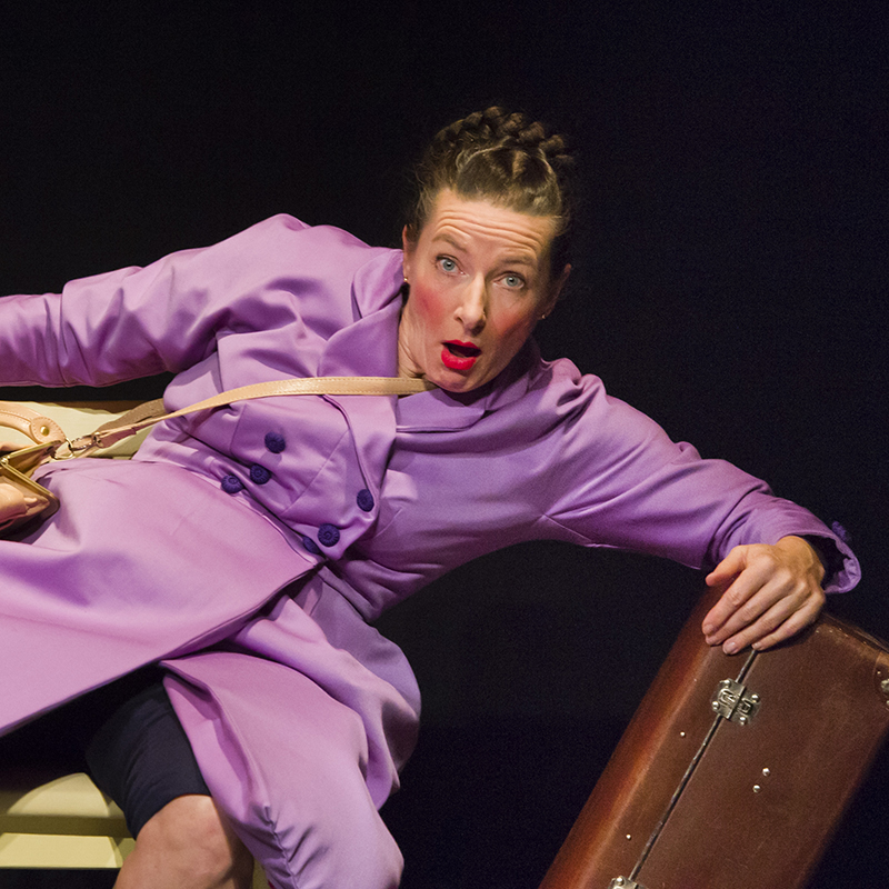 Theaterfoto: Schauspielerin mit langen Zopf, lehnt sich schräg auf einen Koffer und schaut erstaunt