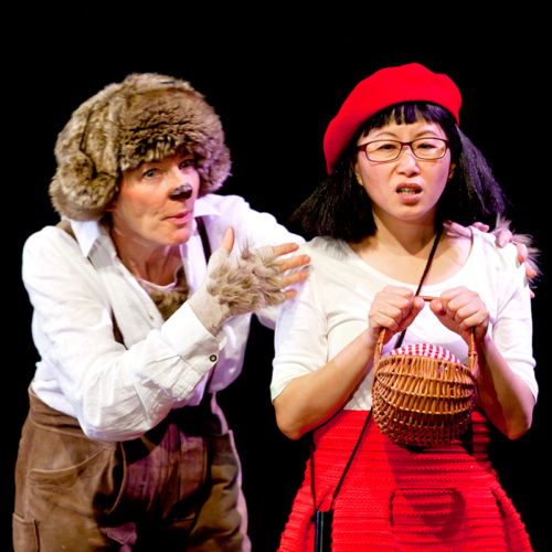 Theaterfoto: Schauspielerin als Wolf verkleidet steht neben Schauspielerin, die mit roter Kappe auf dem Kopf und Körbchen in der Hand als Rotkäppchen verkleidet ist