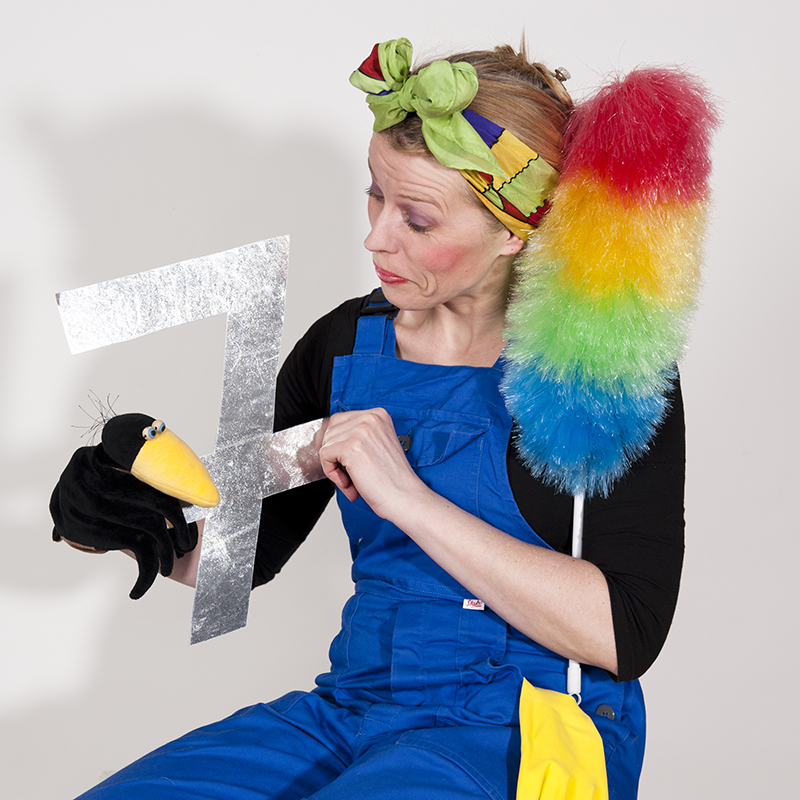 Schauspielerin mit Kopftuch und Staubwedel hält eine Raben-Handpuppe in der Hand und eine Sieben