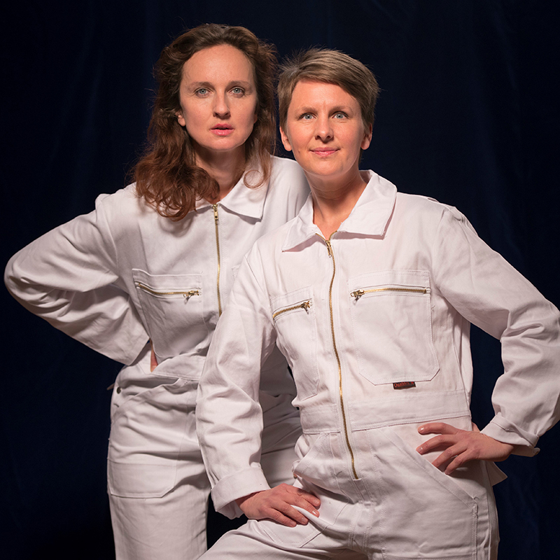 Zwei Schauspielerinnen in weißen Overalls vor schwarzem Hintergrund stehend
