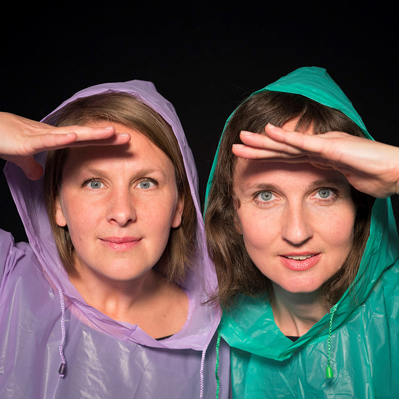 Zwei Schauspielerinnen mit Regenjacken halten sich schützend die Hand über die Stirn