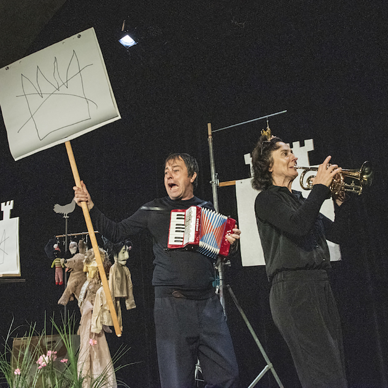 Theaterfoto: Mann mit Akkordeon hält ein Schild mit einer durchgestrichenen Krone nach oben, daneben spielt eine Frau Trompete
